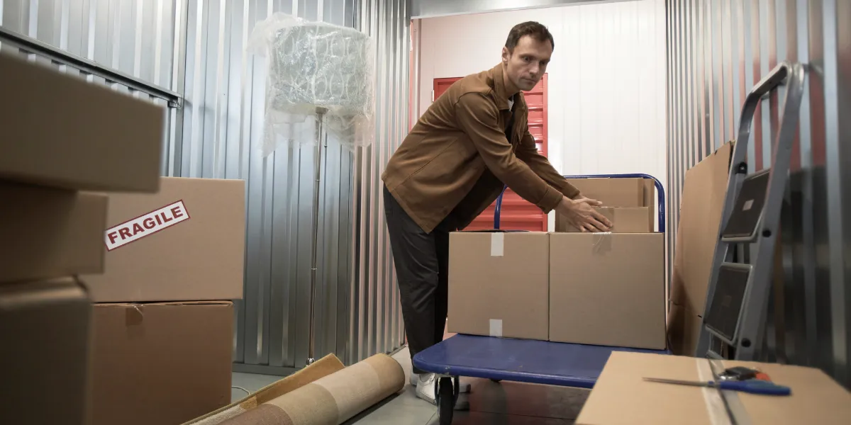 Man packing cartons on a flatbed trolley inside a storage unit, with a box marked fragile beside him