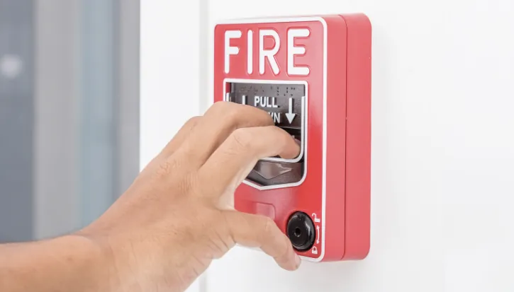Hand pulling a red fire alarm call point mounted on a white wall