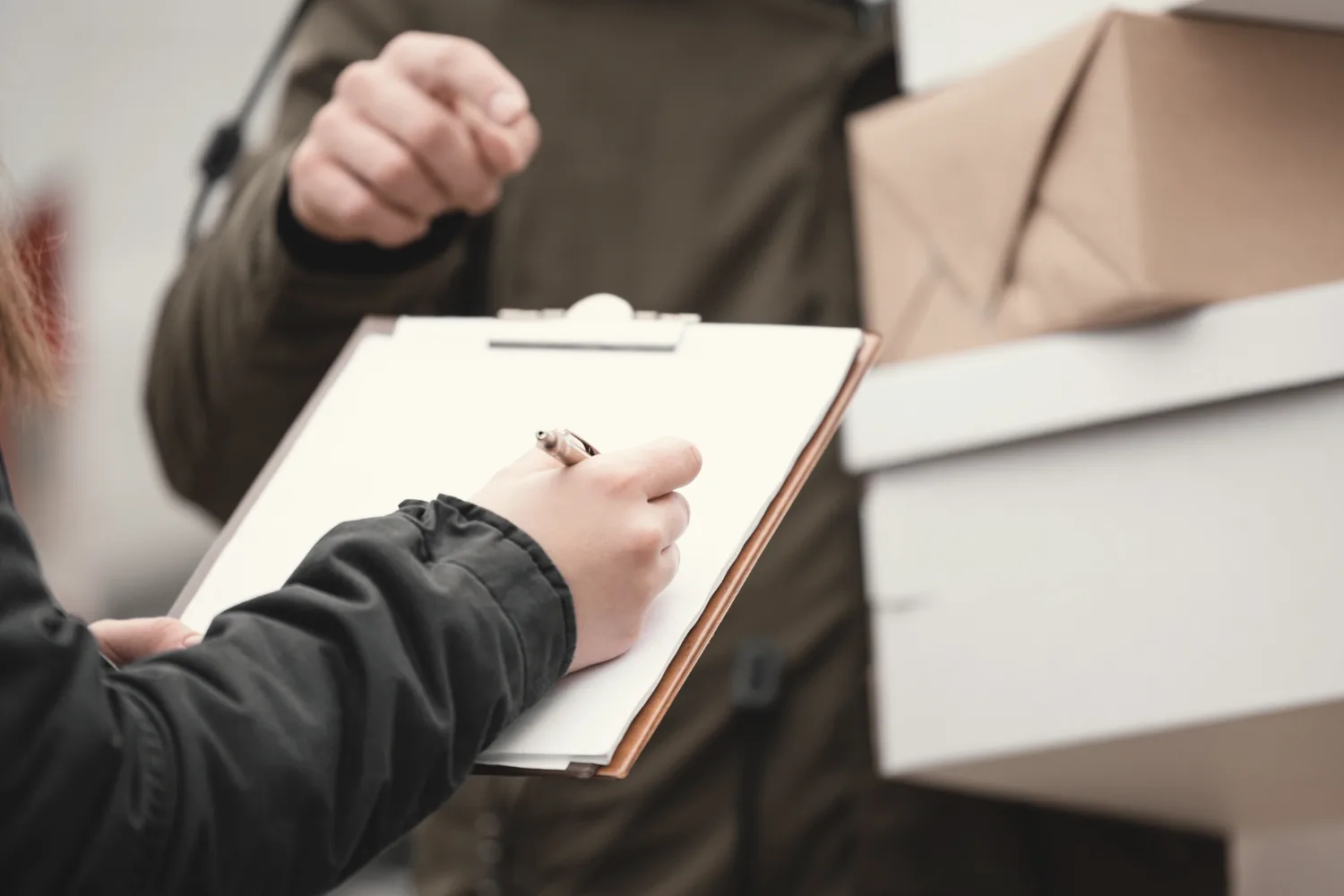 Hand signing a form on a clipboard beside stacked boxes