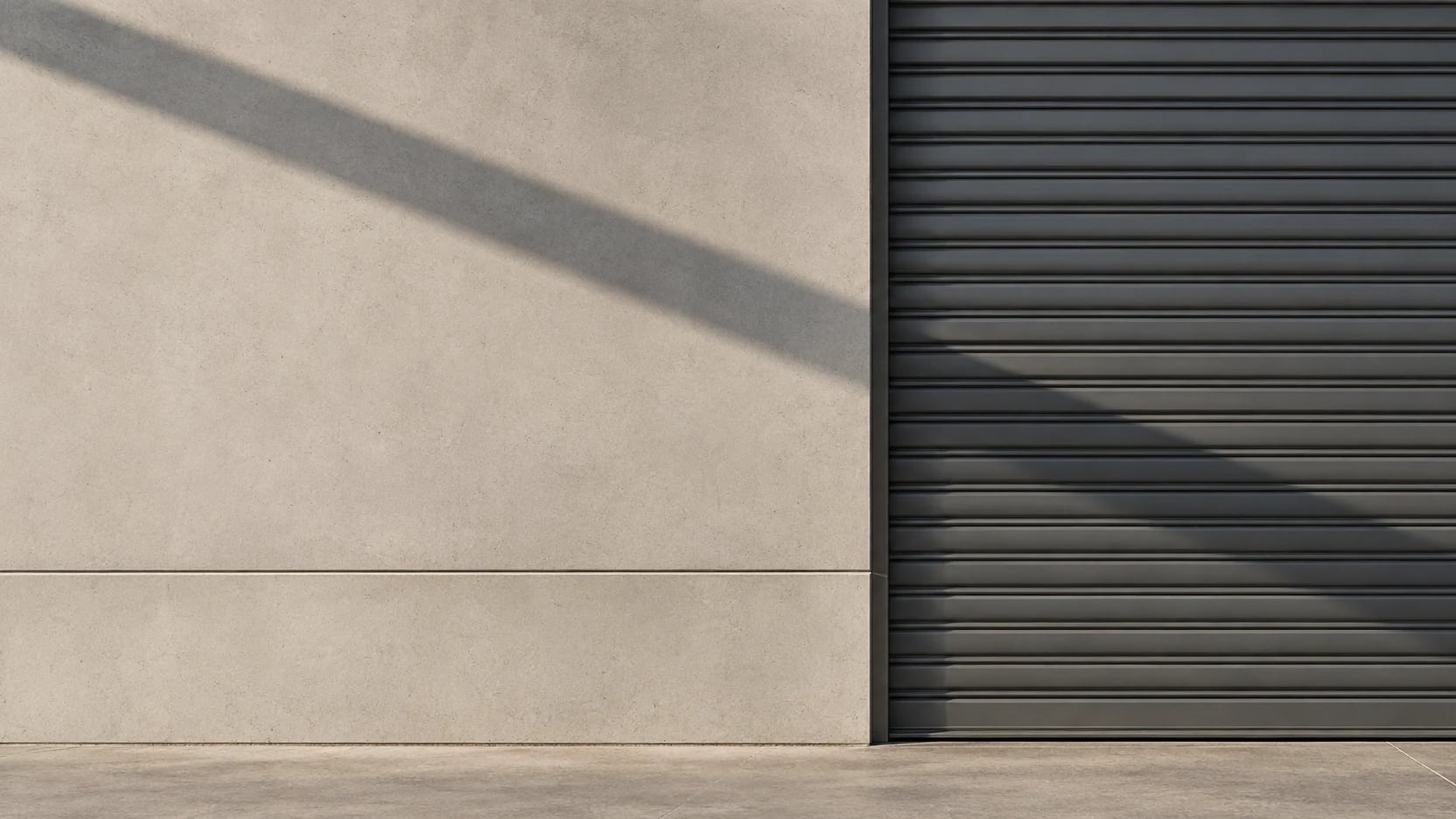 Charcoal roller door set into a pale concrete wall, crossed by a diagonal shadow
